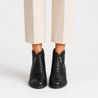 Black leather ankle boots with side zippers, shown on a model wearing beige cropped trousers against a light gray studio background.