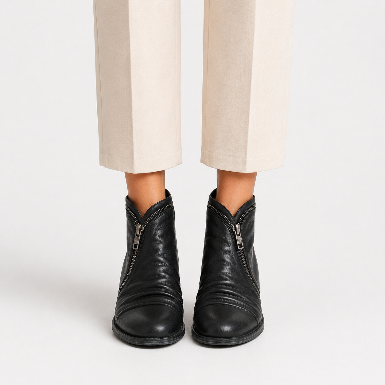 Black leather ankle boots with side zippers, shown on a model wearing beige cropped trousers against a light gray studio background.