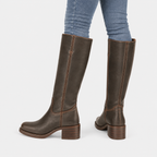 A pair of dark brown leather knee-high boots with stacked block heels, shown on a model wearing blue jeans against a light gray background.