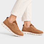 A pair of women's brown knit sneakers with white soles, shown on a model wearing off-white jeans against a light gray studio background.