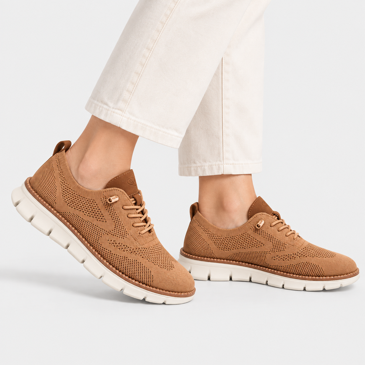 A pair of women's brown knit sneakers with white soles, shown on a model wearing off-white jeans against a light gray studio background.
