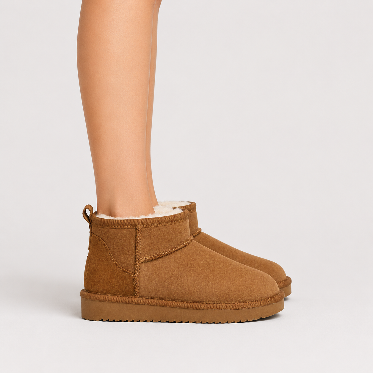 A pair of tan suede ankle boots with white shearling lining, shown on a model's feet against a light gray studio background.