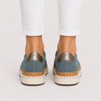 A pair of women's slip-on espadrille sneakers in dusty blue suede with metallic silver heel tabs, cork-wrapped midsoles, and off-white soles, shown on a model wearing white pants from the back against a light gray studio background.