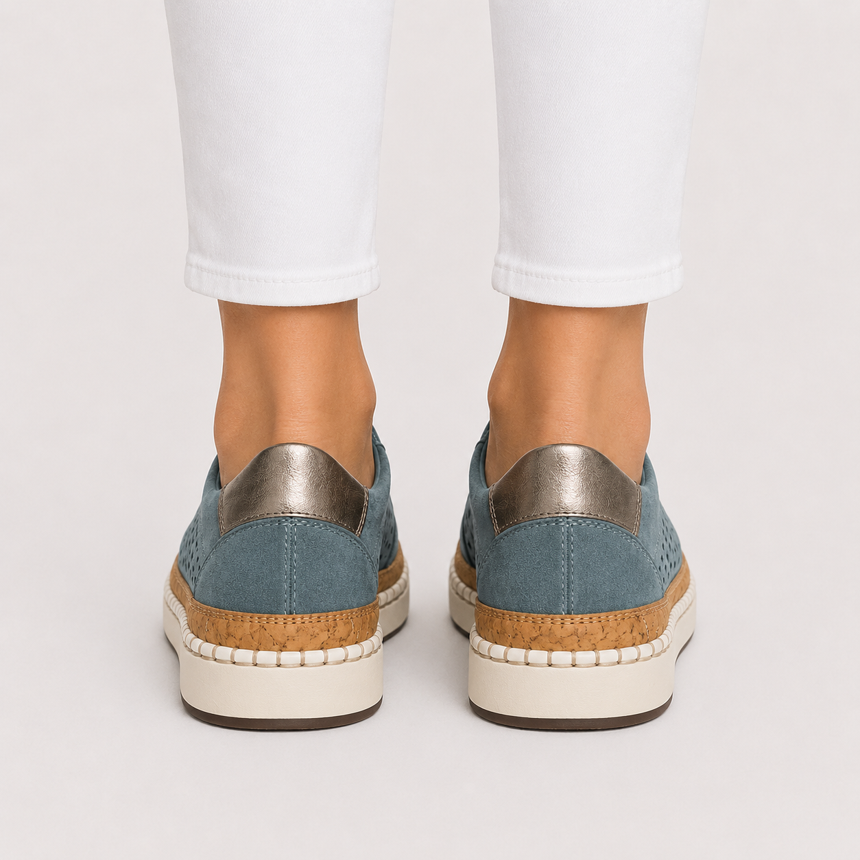 A pair of women's slip-on espadrille sneakers in dusty blue suede with metallic silver heel tabs, cork-wrapped midsoles, and off-white soles, shown on a model wearing white pants from the back against a light gray studio background.