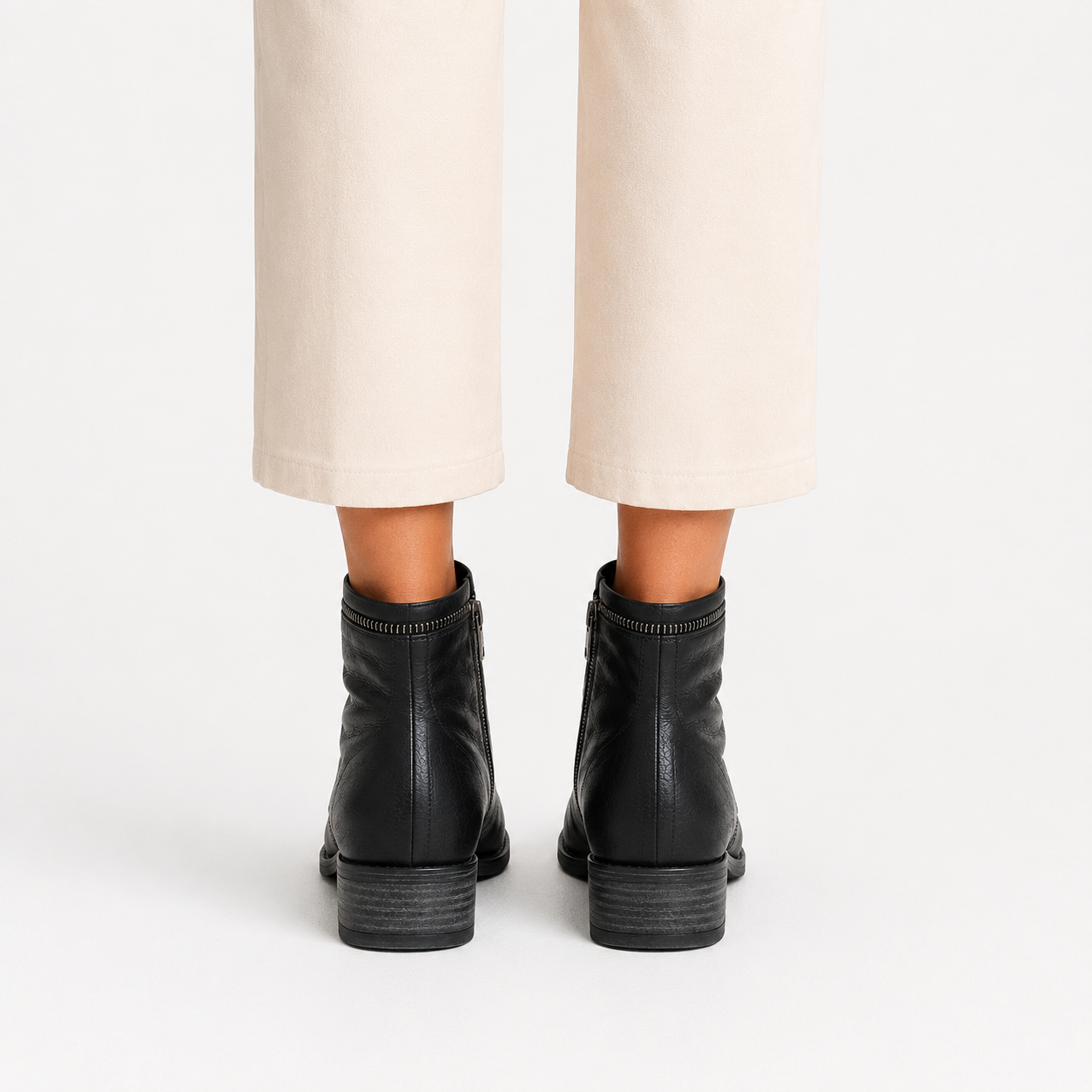 Rear view of a person wearing black leather ankle boots with low block heels and light beige cropped trousers, against a plain white studio background.