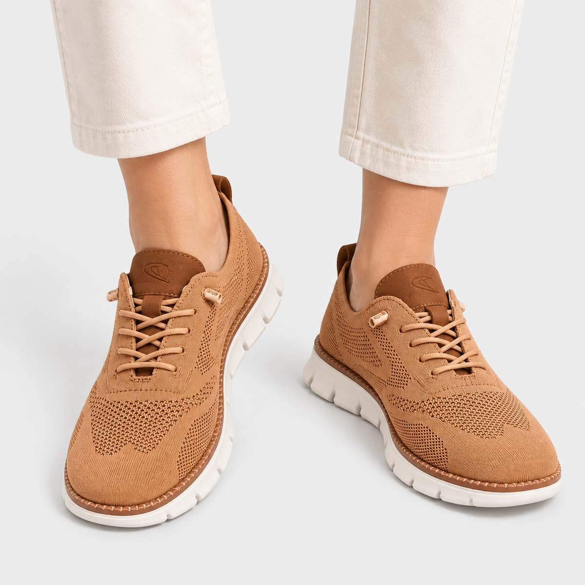 A pair of tan knit lace-up sneakers with white soles, shown on a model wearing off-white pants against a light gray studio background.