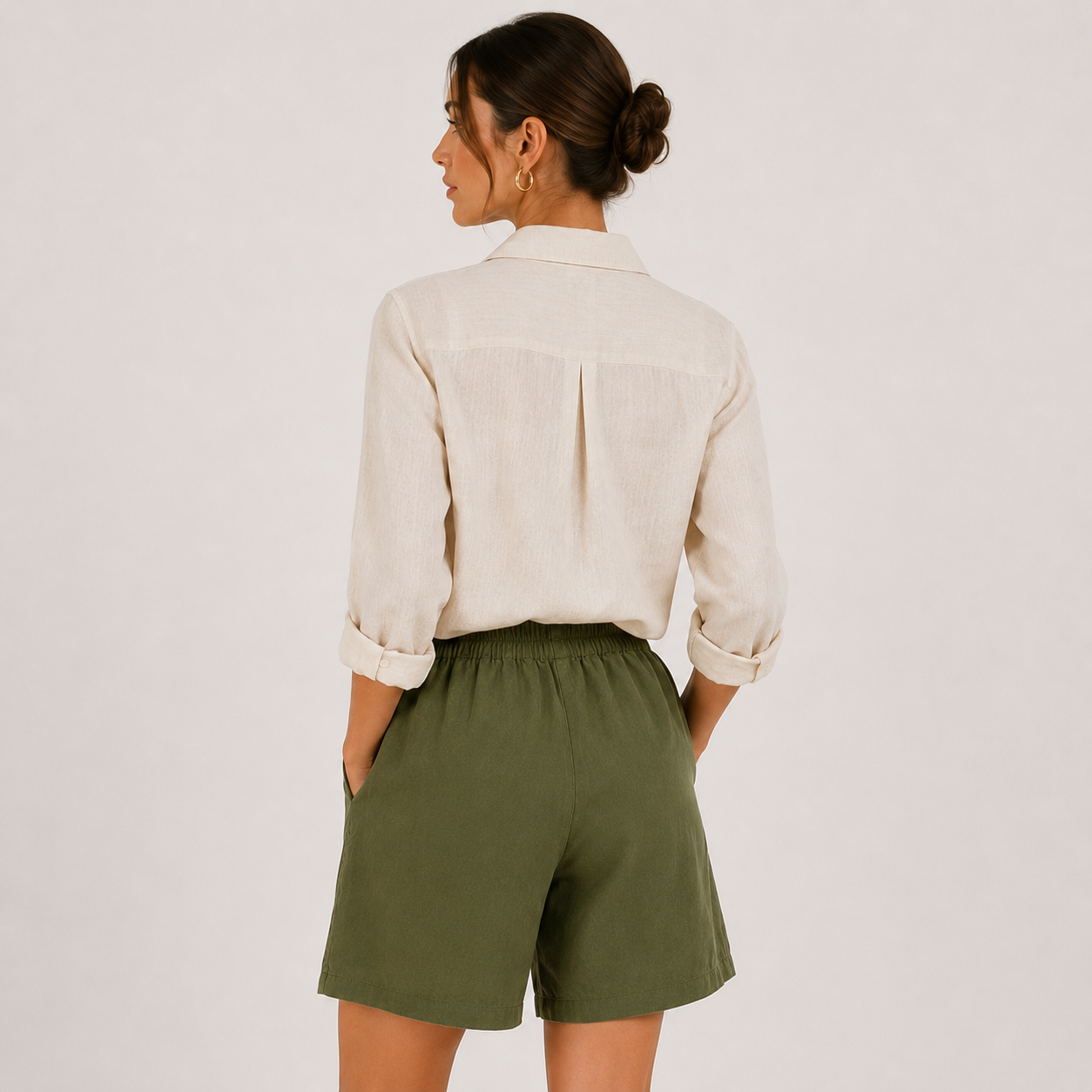 A woman shown from the back wearing a light beige linen button-up shirt and olive green shorts against a plain light gray background.