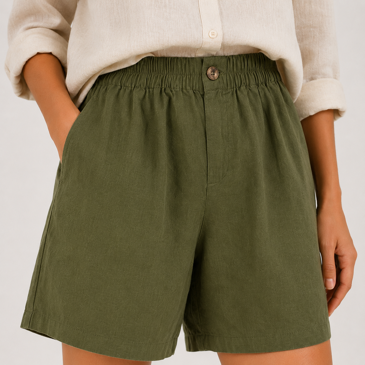 A close-up of a person wearing olive green high-waisted shorts with an elastic waistband and a cream-colored linen button-up shirt.