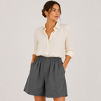 A woman modeling a cream button-up shirt and dark gray high-waisted shorts against a light gray studio background.