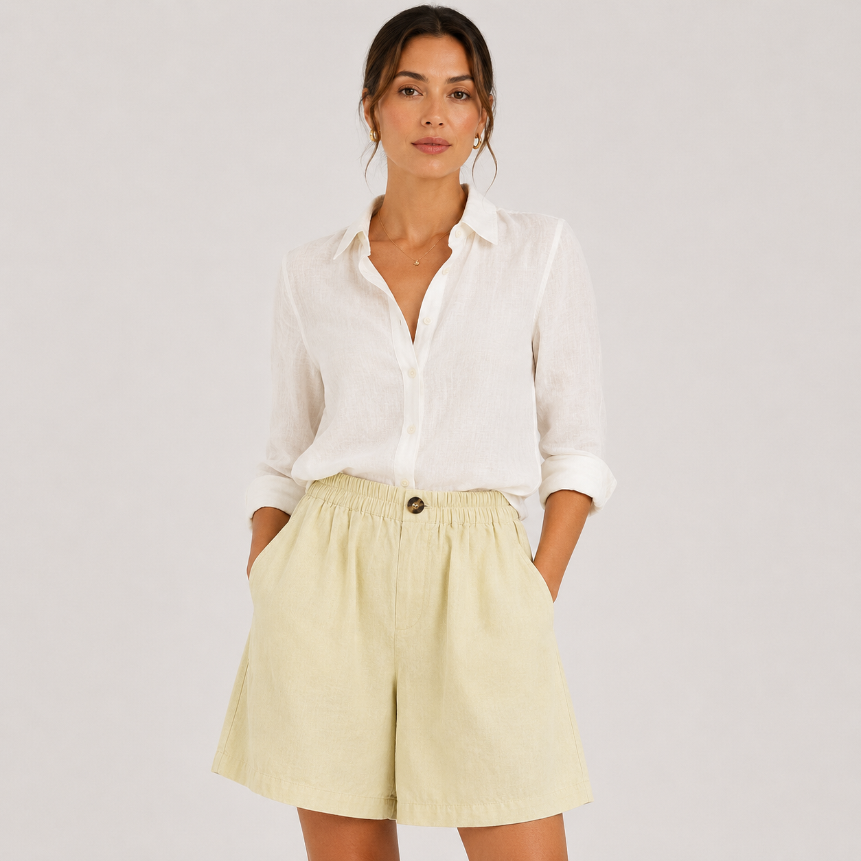 A woman modeling a white button-down shirt and pale yellow high-waisted shorts against a light gray studio background.