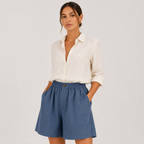 A woman modeling a white linen button-down shirt and blue linen shorts against a light gray studio background.