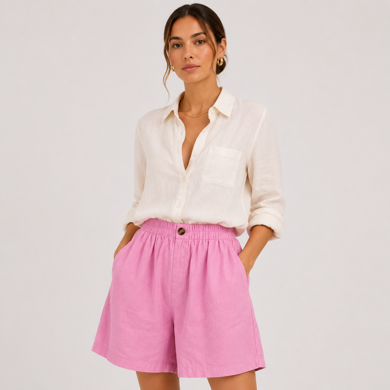 A woman modeling a white button-down shirt and bright pink high-waisted shorts against a light gray studio background.