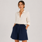A woman modeling a white long-sleeve button-up shirt and navy blue shorts against a light gray studio background.