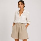 A woman modeling a white long-sleeve button-up shirt and beige linen shorts against a light gray studio background.