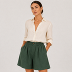 A woman modeling a cream linen button-down shirt and dark green linen shorts against a light gray studio background.