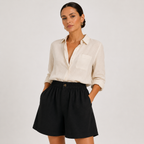 A woman modeling a light cream linen button-down shirt and black high-waisted shorts against a light gray studio background.