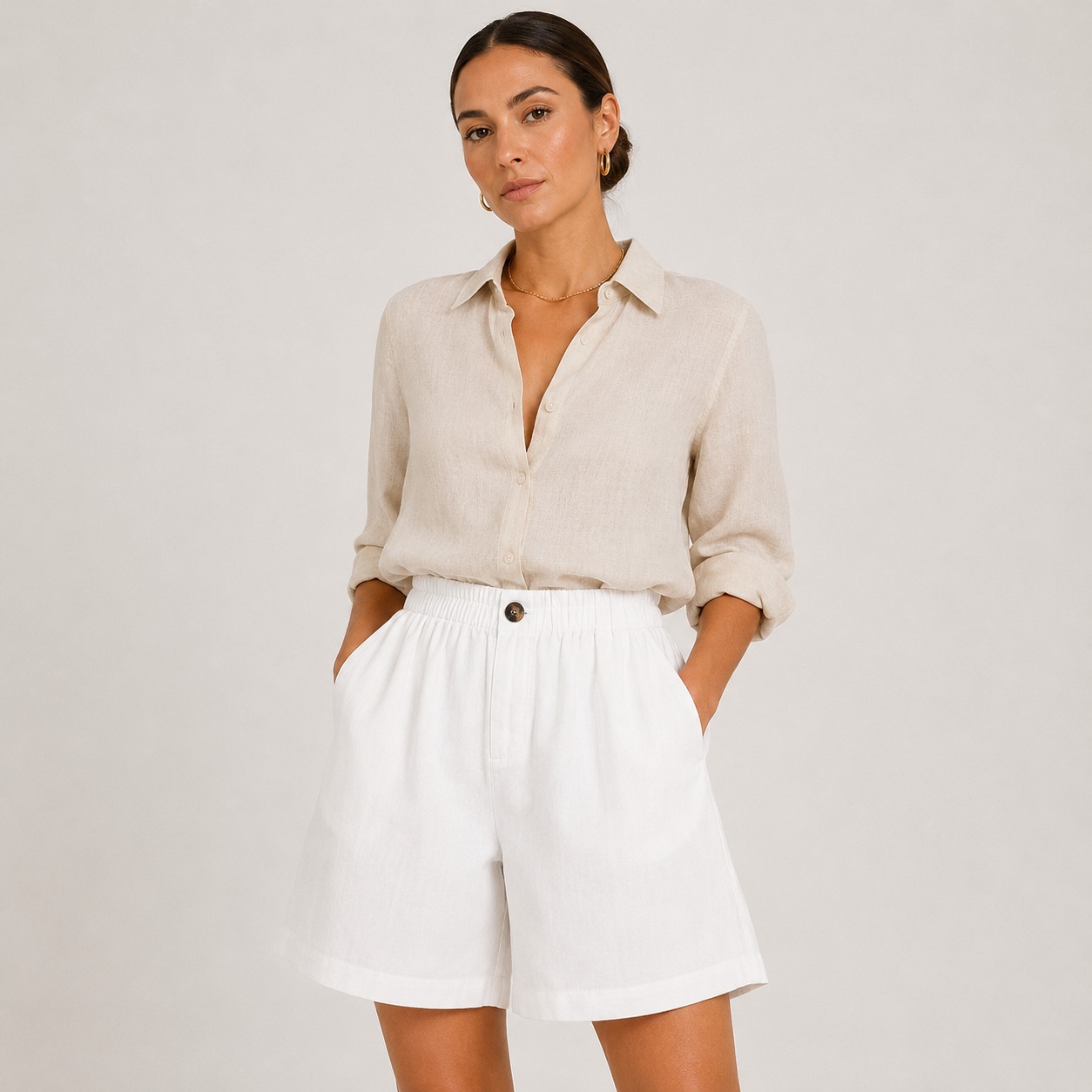 A woman modeling a light beige linen button-down shirt and white shorts against a plain light gray studio background.