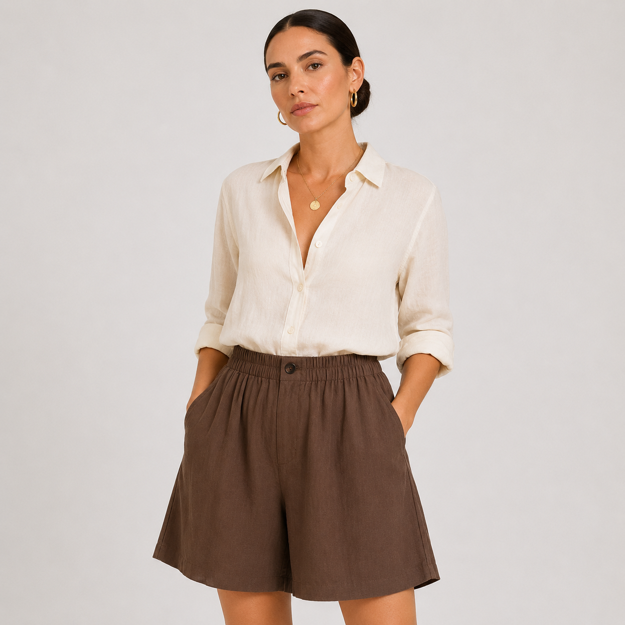 A woman modeling a cream-colored long-sleeve button-down shirt and brown high-waisted shorts against a light gray studio background.