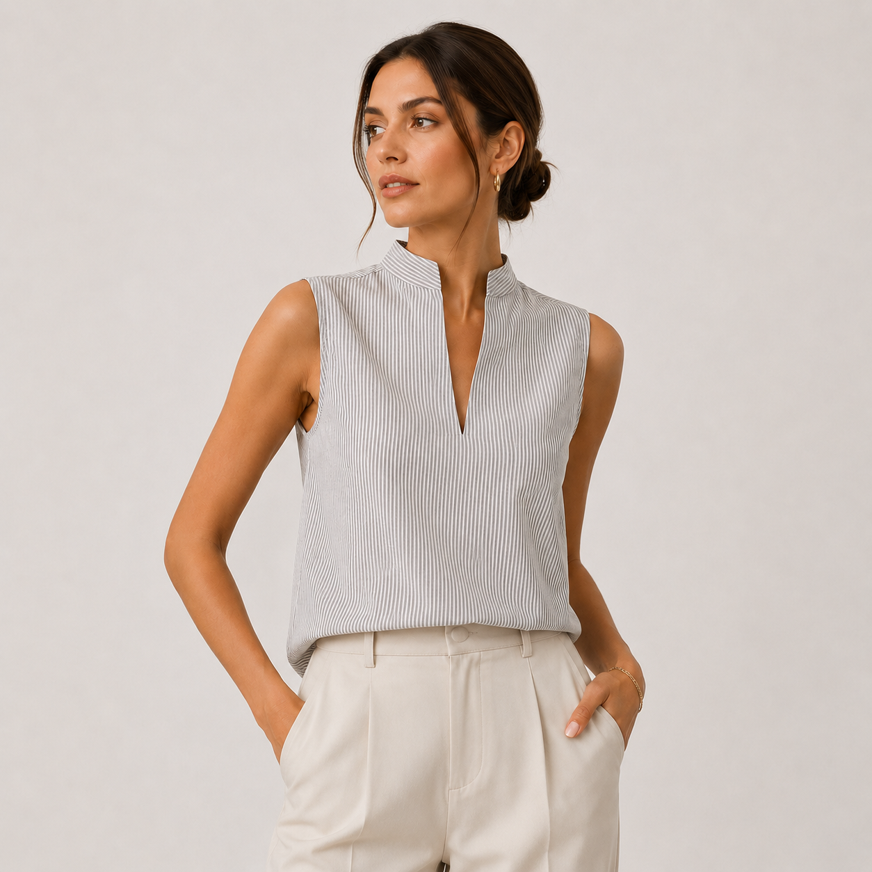 A woman modeling a sleeveless light gray and white vertically striped top with a split V-neckline, paired with cream-colored trousers, against a plain light gray studio background.