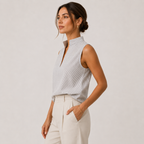 A woman wearing a sleeveless, high-neck top with fine vertical stripes in light gray and white, paired with cream-colored trousers, photographed against a neutral light gray studio background.