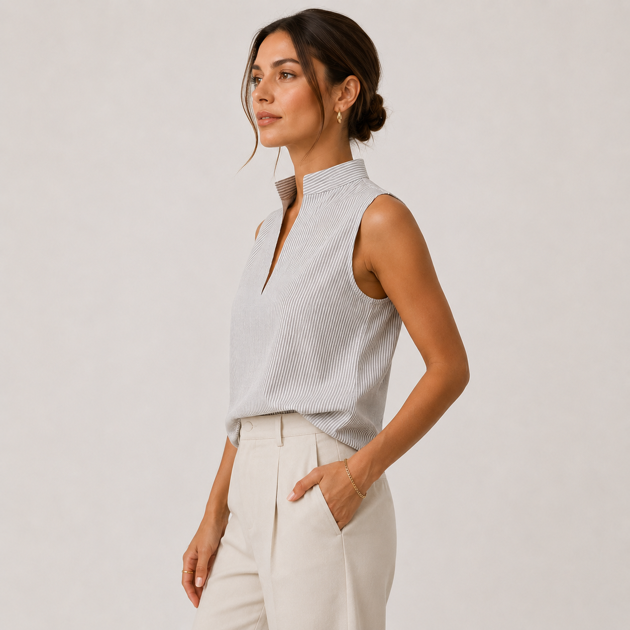 A woman wearing a sleeveless, high-neck top with fine vertical stripes in light gray and white, paired with cream-colored trousers, photographed against a neutral light gray studio background.