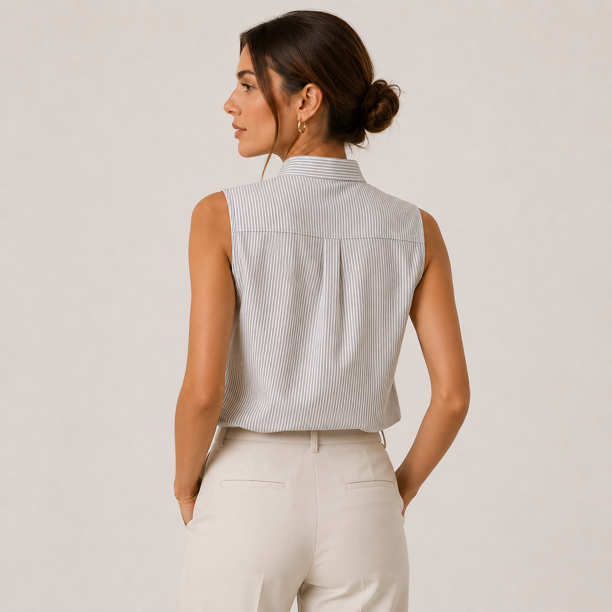 A woman shown from the back wearing a sleeveless, high-neck button-up shirt with fine vertical stripes in light gray and white, paired with cream-colored trousers, against a solid light beige studio background.