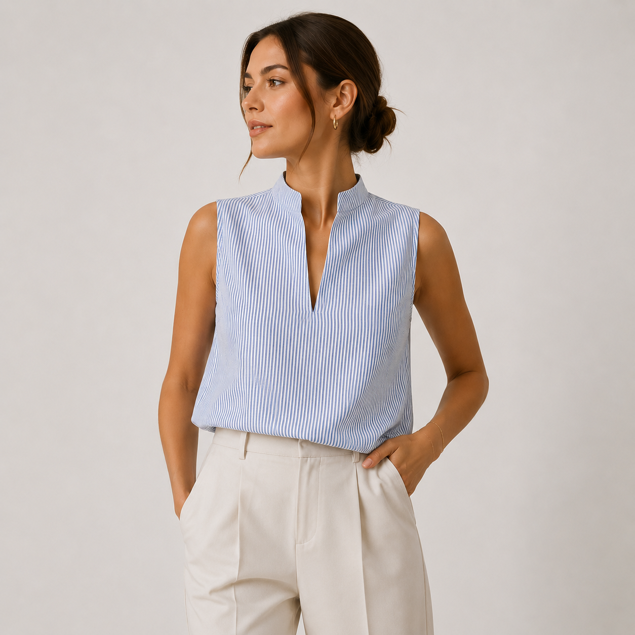 A woman wearing a sleeveless light blue and white striped top with a split V-neckline, paired with cream-colored trousers, standing against a neutral light gray studio background.