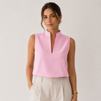 A woman wearing a sleeveless pink and white vertically striped top with a mock neck and split V-neckline, paired with light beige trousers, against a plain light gray studio background.