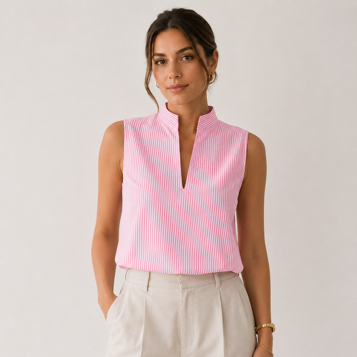 A woman wearing a sleeveless pink and white vertically striped top with a mock neck and split V-neckline, paired with light beige trousers, against a plain light gray studio background.