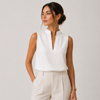 A woman wearing a white sleeveless ribbed blouse with a split neckline and cream-colored trousers, standing against a light gray studio background.