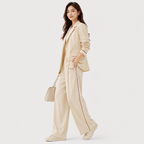 A woman models a matching beige two-piece lounge set consisting of a long-sleeve button-front blazer and wide-leg trousers with thin dark piping details, paired with a matching beige handbag and beige sneakers against a light gray studio background.