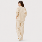 A woman is shown from behind wearing a light beige two-piece lounge set consisting of a long-sleeve top and matching wide-leg pants, set against a plain light gray studio background.