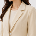 A close-up of a woman wearing a light beige blazer over a matching beige top, showing the collar, lapels, and a single visible button against a plain light background.