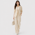 A woman modeling a light beige two-piece suit consisting of a blazer and wide-leg trousers, paired with a white top, beige sneakers, and a matching beige handbag against a light gray studio background.