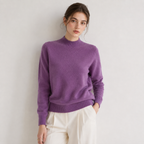 A woman wearing a purple knit mock neck sweater and cream-colored pants, standing against a light gray wall.