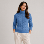 A woman wearing a blue cable-knit turtleneck sweater and cream-colored trousers, standing against a neutral light gray studio background.