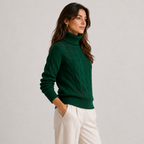 A woman wearing a dark green cable-knit turtleneck sweater and off-white trousers, standing against a light gray studio background.