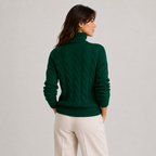 A woman shown from the back wearing a dark green cable-knit turtleneck sweater and cream-colored trousers, standing against a light gray studio background.