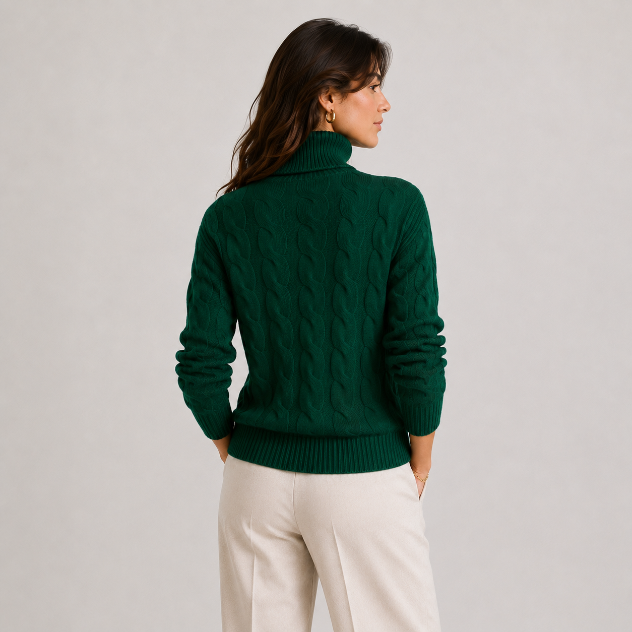 A woman shown from the back wearing a dark green cable-knit turtleneck sweater and cream-colored trousers, standing against a light gray studio background.