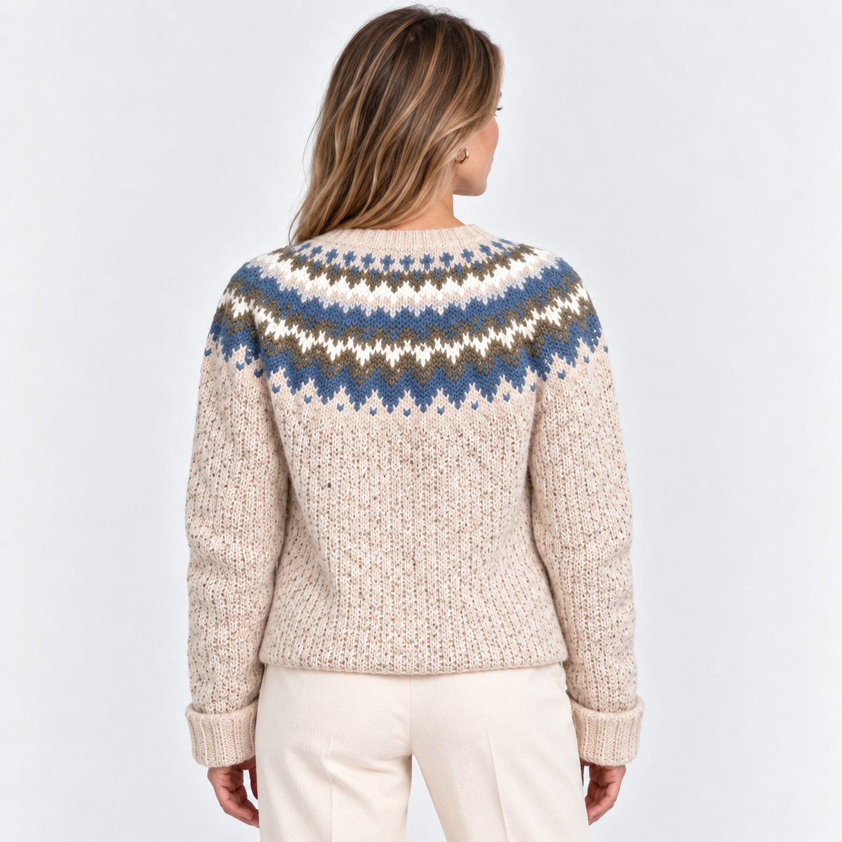 A woman viewed from the back wearing a cream-colored knit sweater with a blue, brown, and white Fair Isle yoke pattern, paired with cream pants, against a light gray studio background.