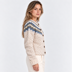 A woman wearing a cream-colored knit cardigan with a blue, brown, and white Fair Isle pattern across the yoke, paired with cream trousers, photographed against a light gray studio background.