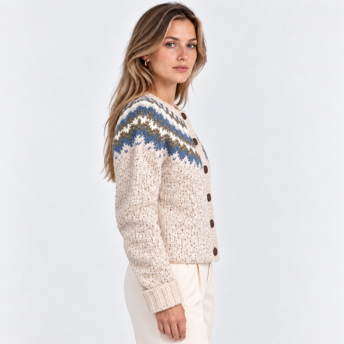 A woman wearing a cream-colored knit cardigan with a blue, brown, and white Fair Isle pattern across the yoke, paired with cream trousers, photographed against a light gray studio background.