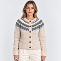A woman wearing a cream-colored knit cardigan with a blue and brown Fair Isle pattern across the yoke and brown buttons, paired with cream trousers, against a light gray studio background.