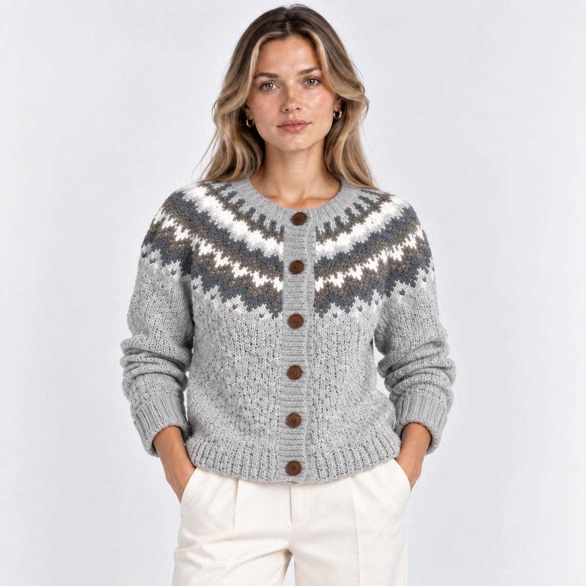 A woman modeling a light gray knit cardigan with a dark gray and white Fair Isle pattern across the chest and waist, featuring brown buttons, paired with cream trousers against a light gray studio background.