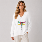 A woman wearing a white knit V-neck sweater with a colorful watercolor dragonfly graphic on the front, paired with light beige pants, against a plain light gray studio background.