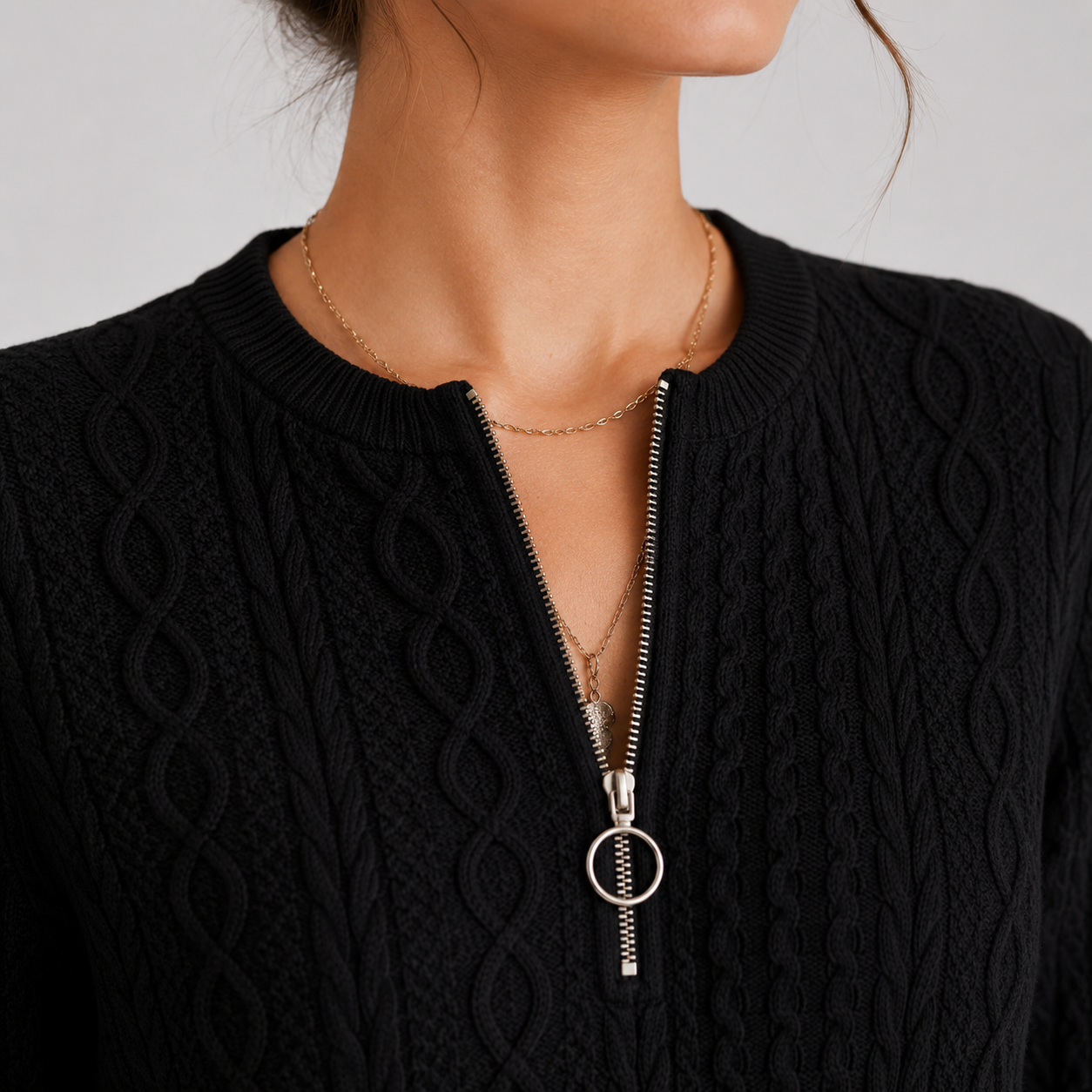 Close-up of a woman wearing a black cable-knit zip-up sweater with a silver-tone zipper and circular ring pull, layered with delicate gold necklaces.