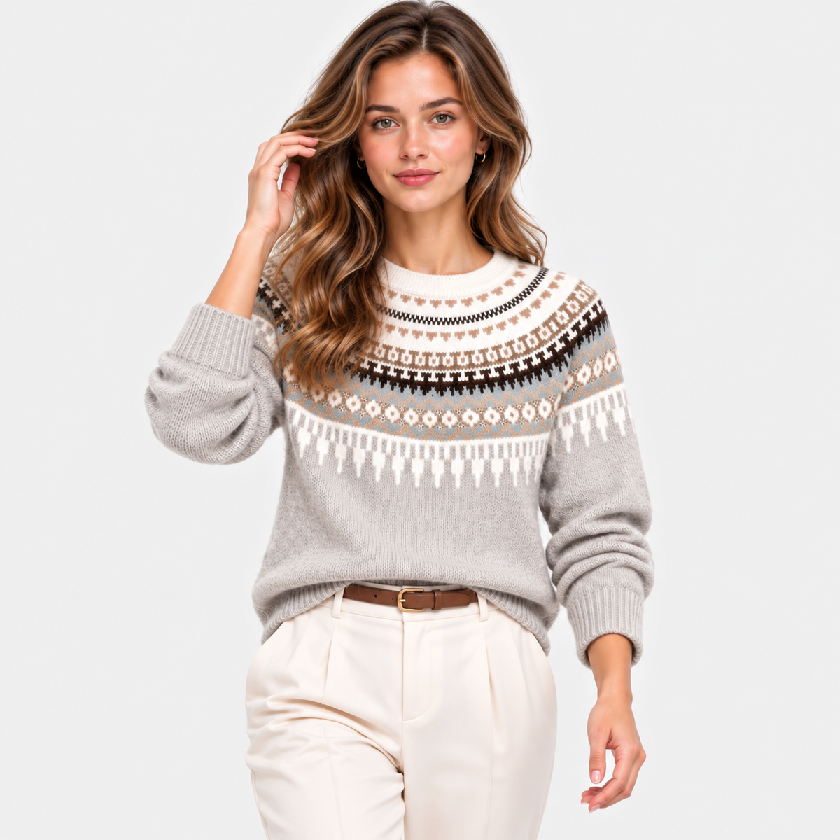 A woman modeling a light gray knit sweater with a traditional Nordic-style pattern in cream, brown, and black, paired with cream trousers, against a solid light gray background.