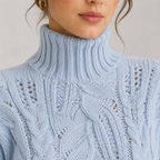 A close-up of a woman wearing a light blue cable-knit turtleneck sweater, showing the detailed knit pattern and her lower face.