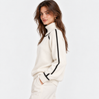 A woman wearing a cream-colored knit turtleneck sweater with black stripe accents down the sleeves, paired with matching cream pants, set against a plain white studio background.
