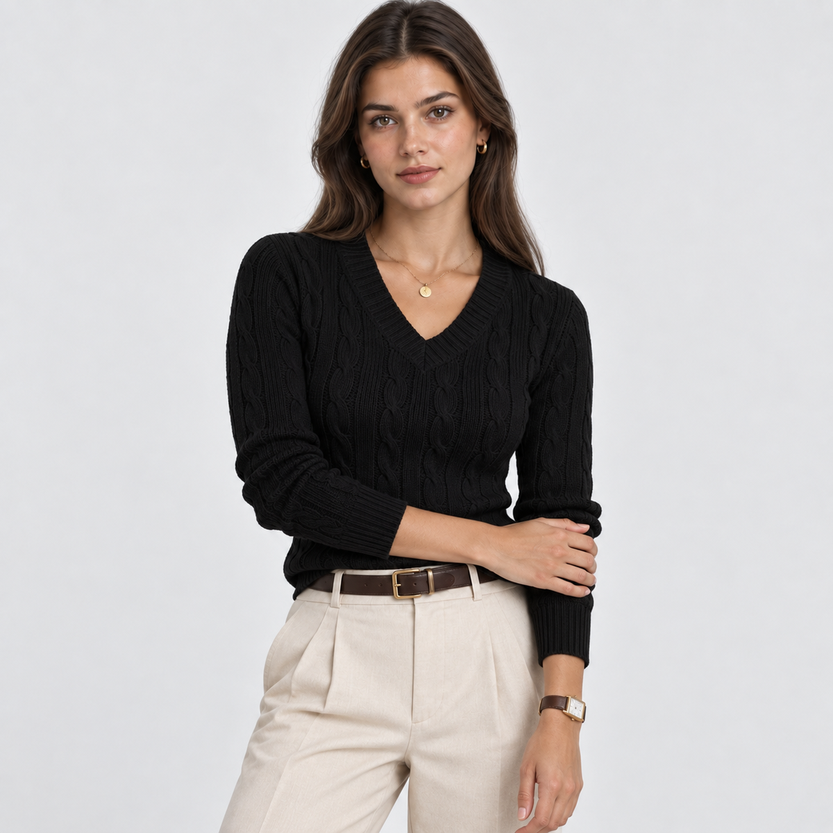 A woman modeling a black cable-knit V-neck sweater with cream-colored trousers and a brown belt, photographed against a light gray studio background.